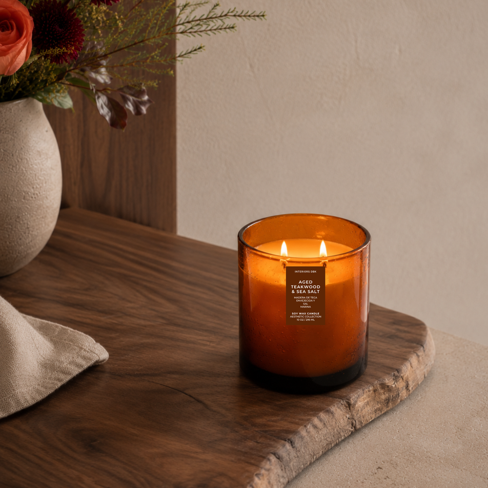 Aged Teakwood and Salt soy wax candle in a amber glass vessel sits on a rustic wooden board in a styled interior setting. The candle features two lit wicks.