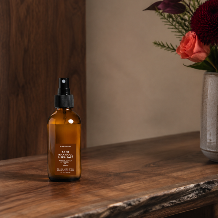 Aged Teakwood and Sea Salt room spray in amber glass bottle placed on a rustic wooden table next to a vase of flowers against a warm neutral wall.