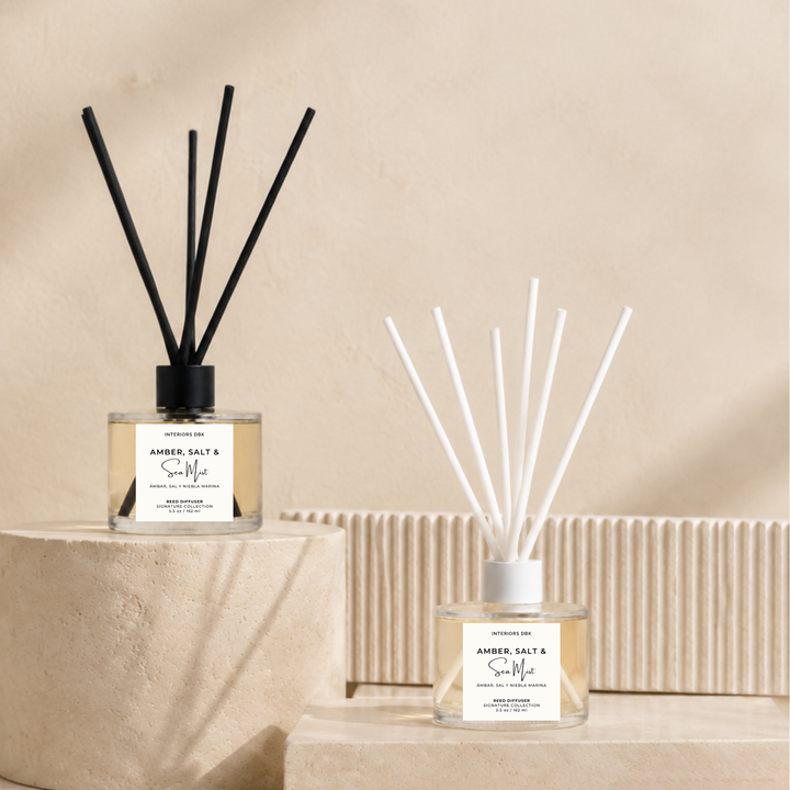 Two amber salt &amp; sea moss reed diffusers styled on beige stone pedestals against a textured beige wall. One diffuser features black reeds and the other features white reeds, both in clear glass bottles with amber liquid and white labels.