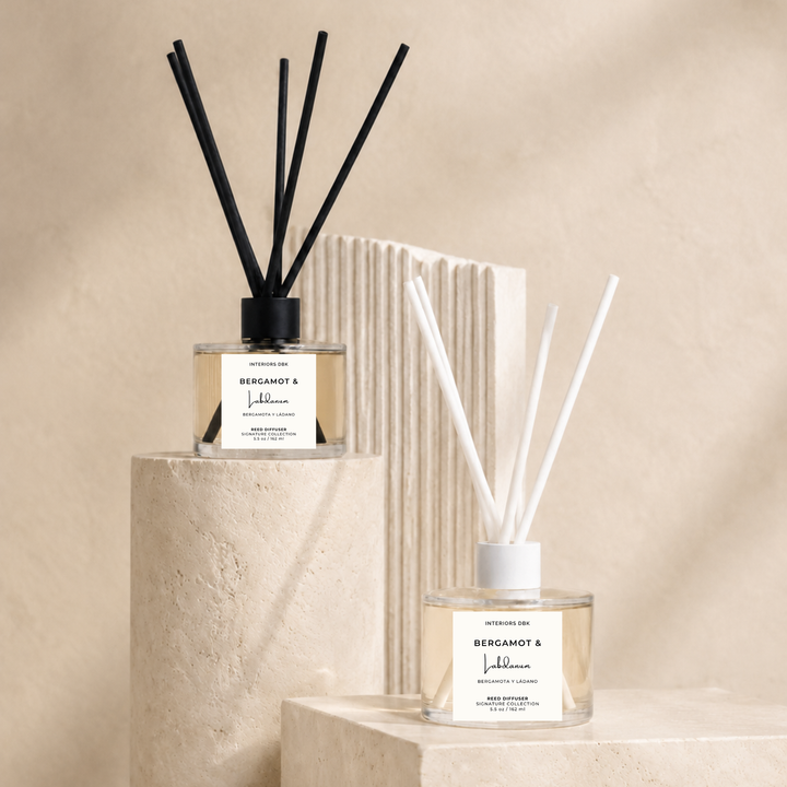 Two Bergamot and Labdanum reed diffusers by Interiors DBK displayed on a beige stone pedestal against a textured beige wall. One diffuser features black reeds in a clear glass bottle with a black cap, and the other features white reeds in a matching bottle with a white cap.