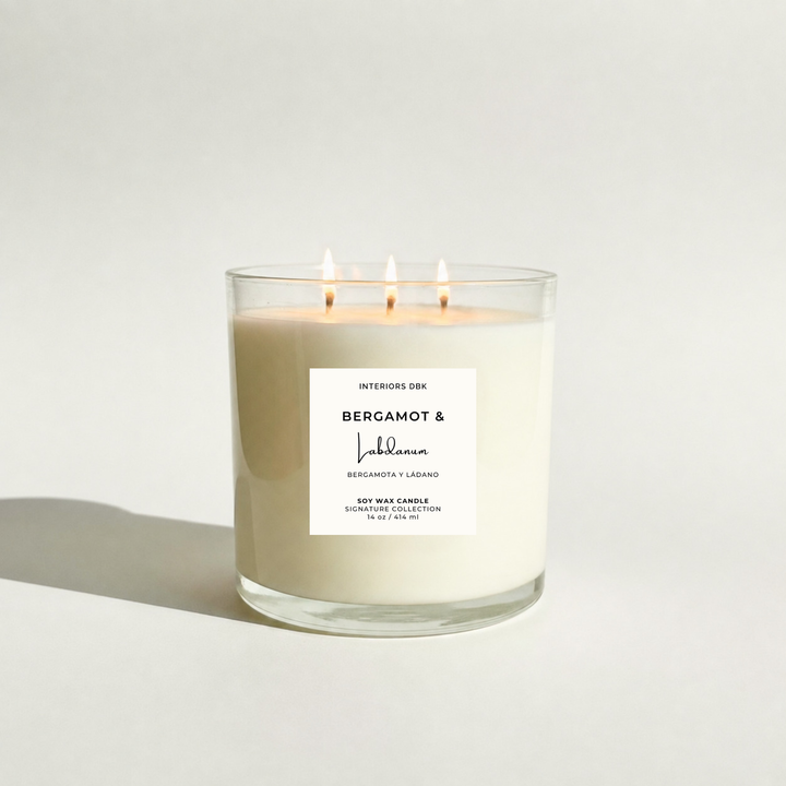 A three-wick soy wax candle in a clear glass jar with a product label reading Bergamot and Labdanum by Interiors DBK, lit with three flames against a clean white background.