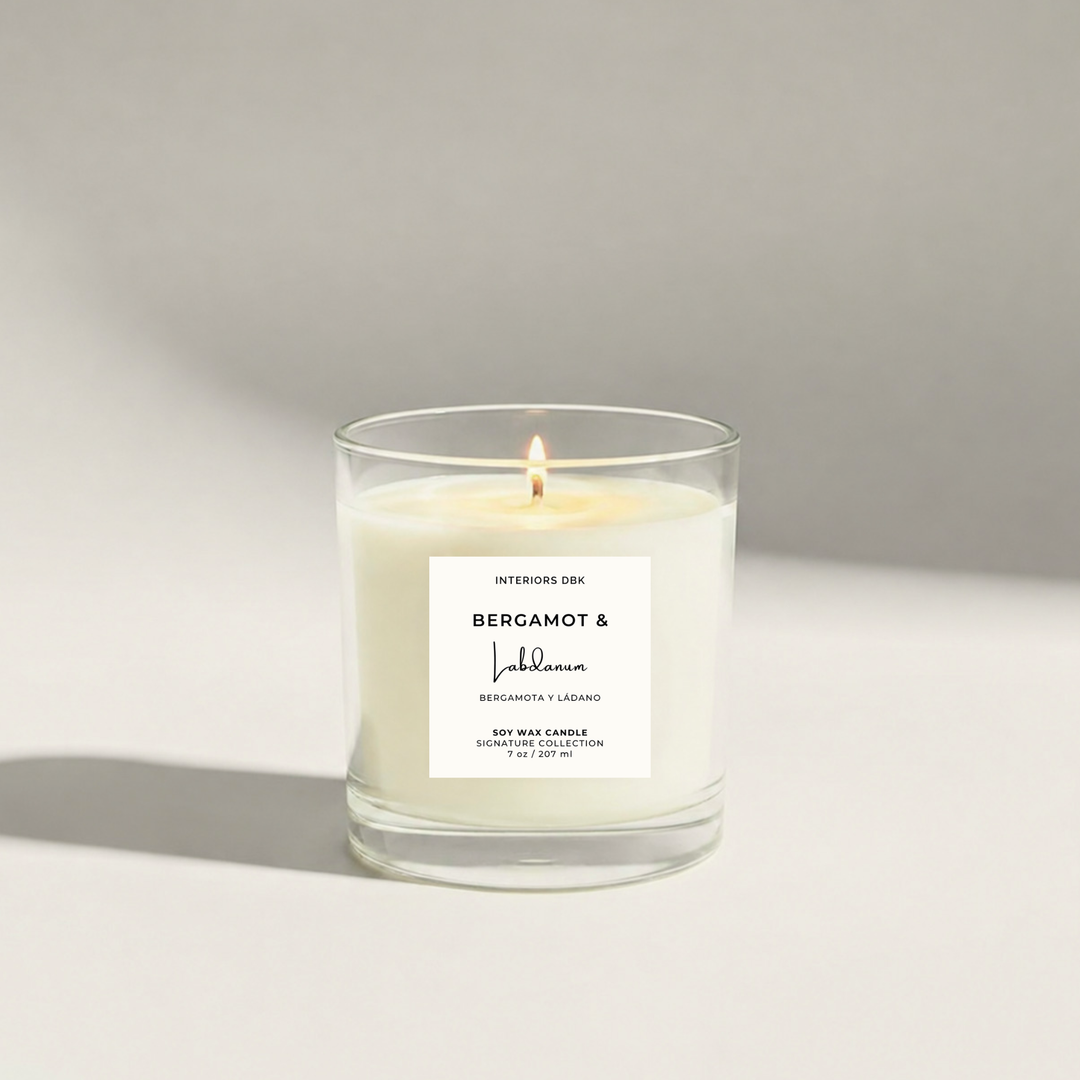 A lit scented soy wax candle in a clear glass jar with a product label reading Bergamot &amp; Labdanum 6.5 oz soy candle by Interiors DBK, set against a minimalist white background with a soft shadow.