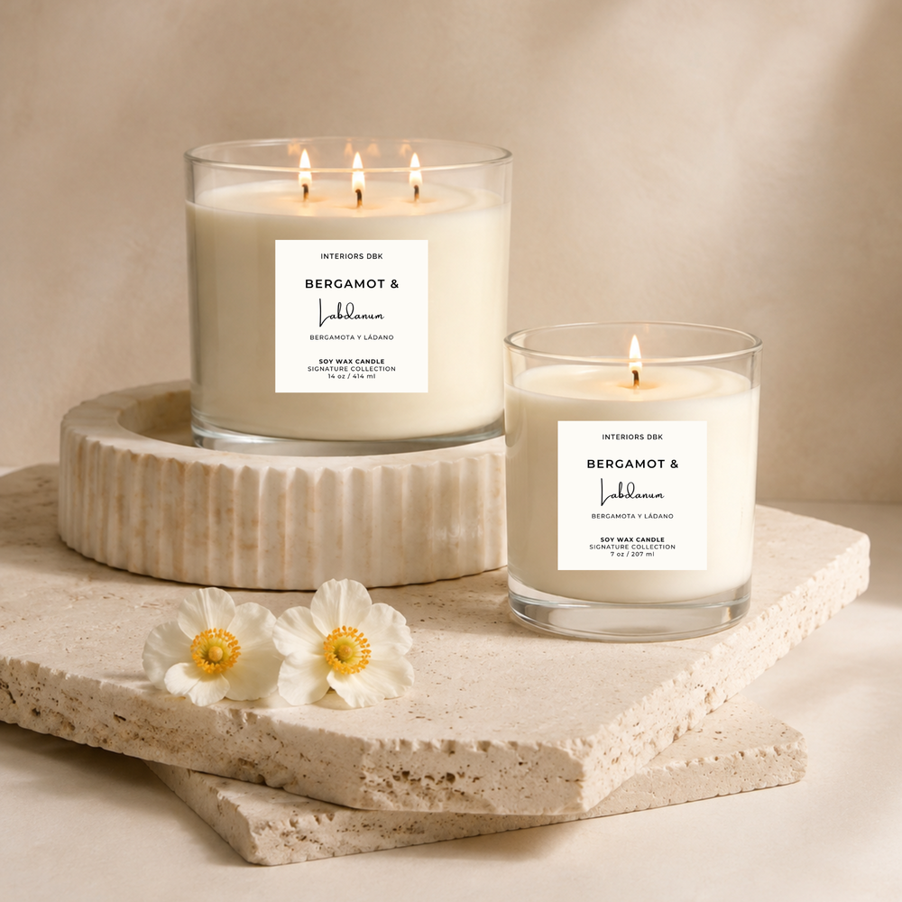Two luxury home fragrances. Soy wax candles with white labels reading Bergamot and Labdanum by Interiors DBK, styled on stacked stone slabs with white flowers against a warm light beige background.