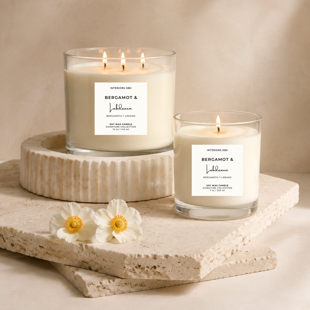 Two luxury home fragrances. Soy wax candles with white labels reading Bergamot and Labdanum by Interiors DBK, styled on stacked stone slabs with white flowers against a warm light beige background.