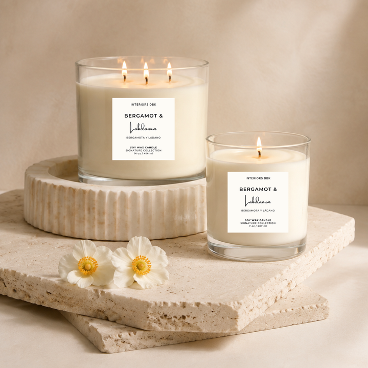 Two luxury home fragrances. Soy wax candles with white labels reading Bergamot and Labdanum by Interiors DBK, styled on stacked stone slabs with white flowers against a warm light beige background.