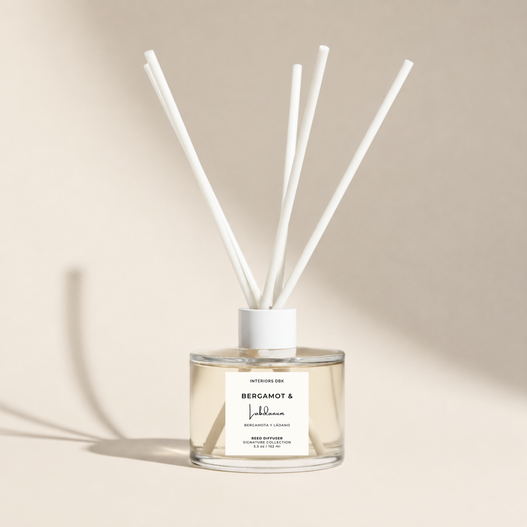 A clear glass reed diffuser bottle filled with diffuser oil and white fiber reeds extending upward. A product label on the front reads Bergamot and Labdanum, a luxury home fragrance by Interiors DBK.
