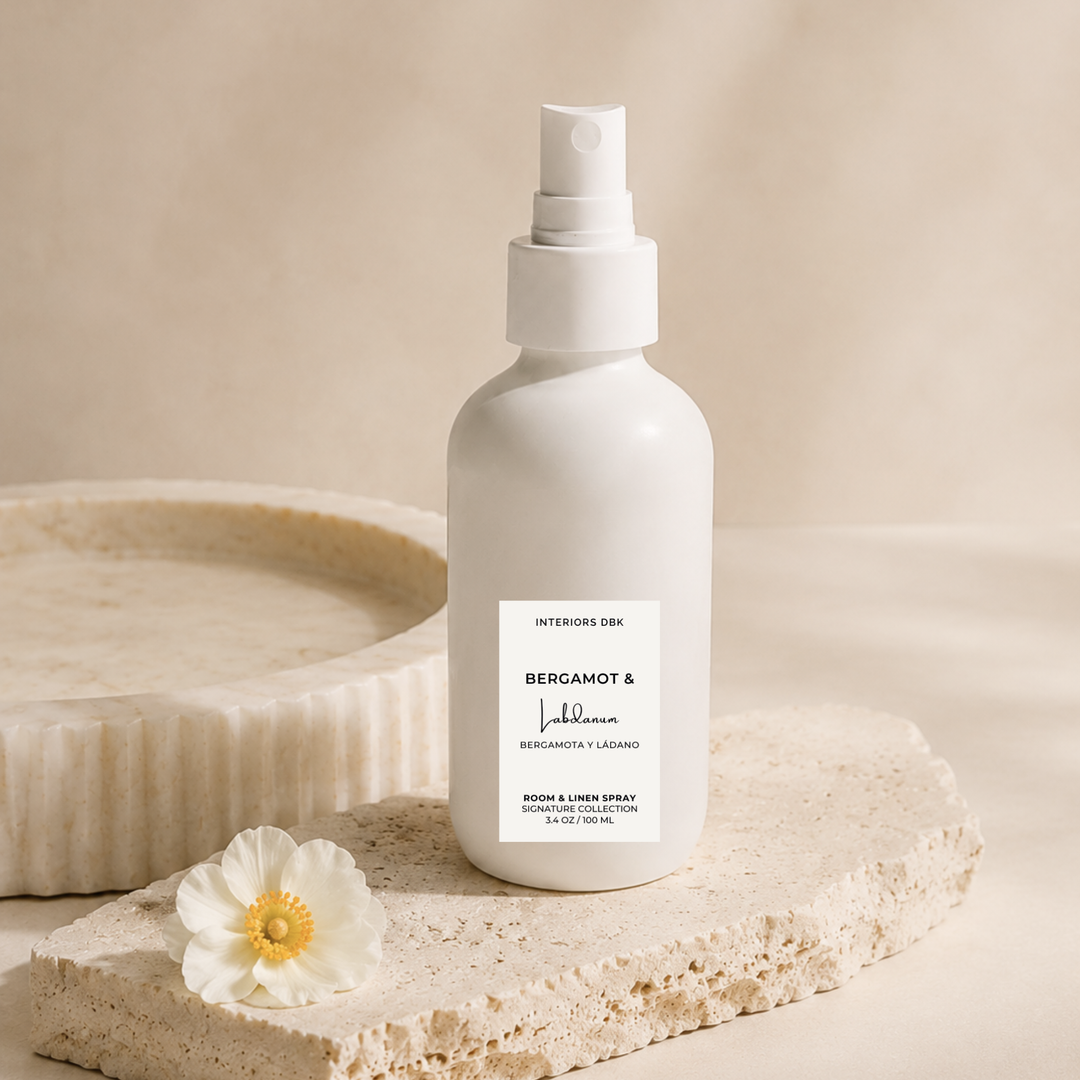 A room and linen spray bottle with a white pump, standing on a textured stone slab next to a white flower. The bottle label reads Bergamot and Labdanum by Interiors DBK.