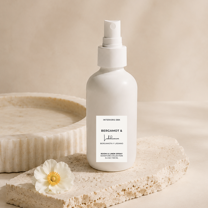 A room and linen spray bottle with a white pump, standing on a textured stone slab next to a white flower. The bottle label reads Bergamot and Labdanum by Interiors DBK.