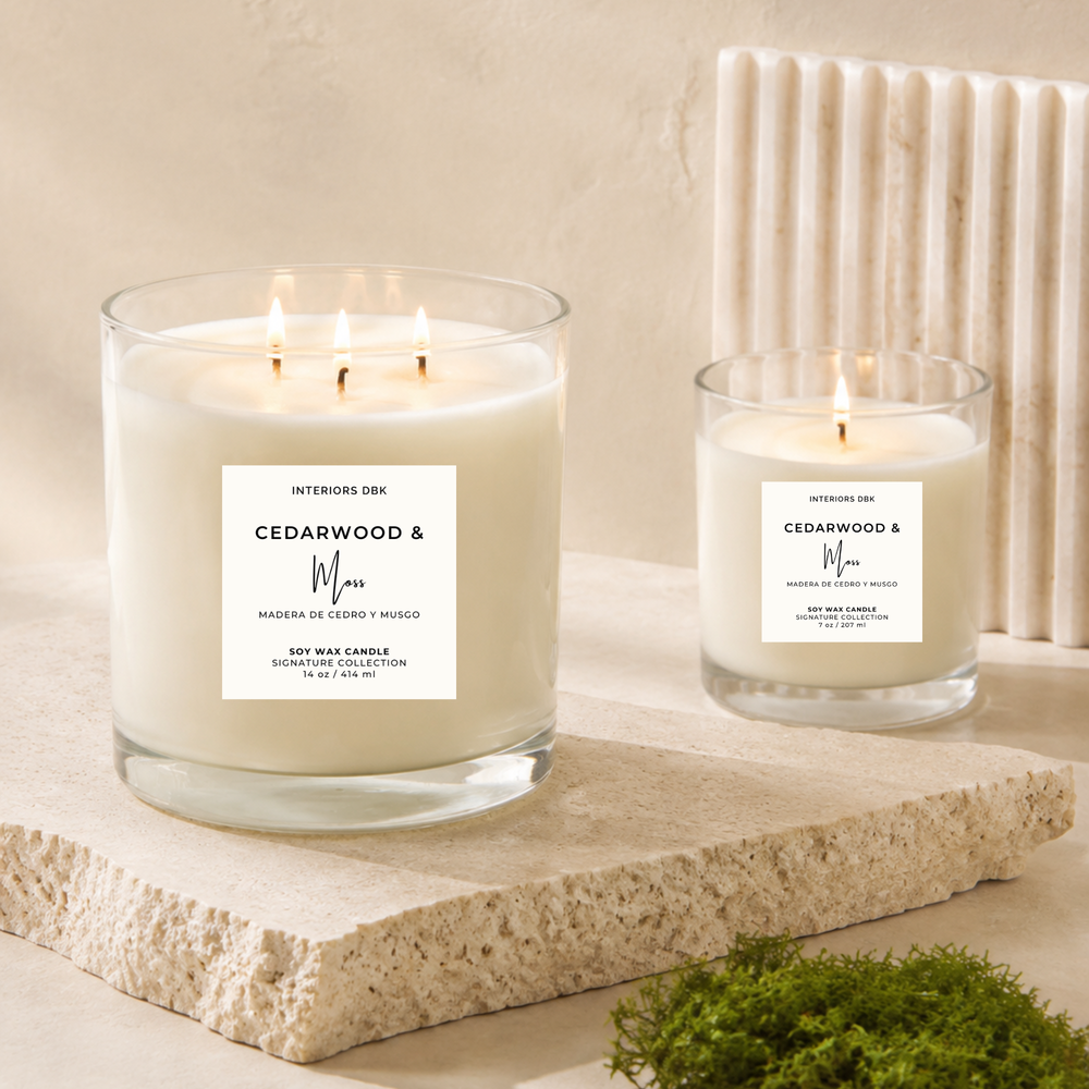 Cedarwood and moss soy wax candles by Interiors DBK. This image displays two lit soy wax candles in clear glass jars photographed on a clean light beige studio background.