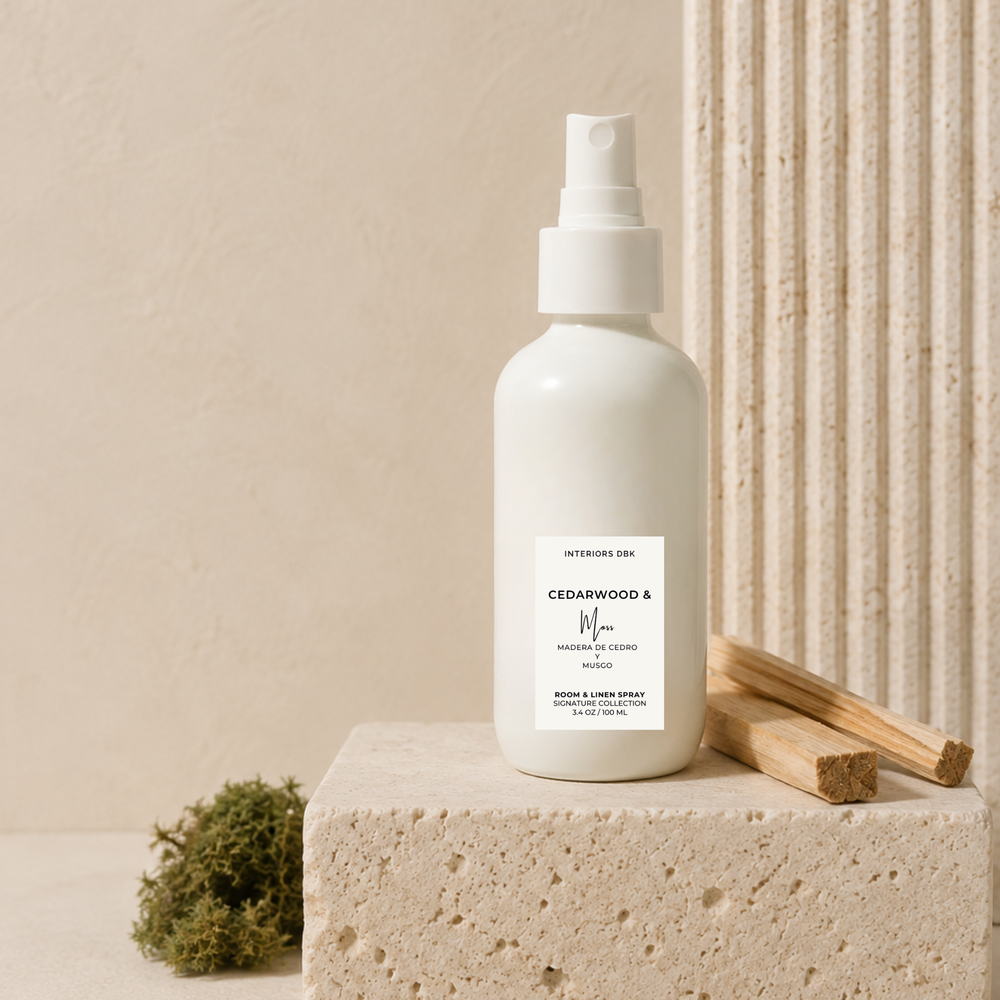 Cedarwood and moss room and linen spray by Interiors DBK. A white spray bottle stands on a textured stone block, styled with a small green moss sprig and wooden incense sticks against a warm beige background.
