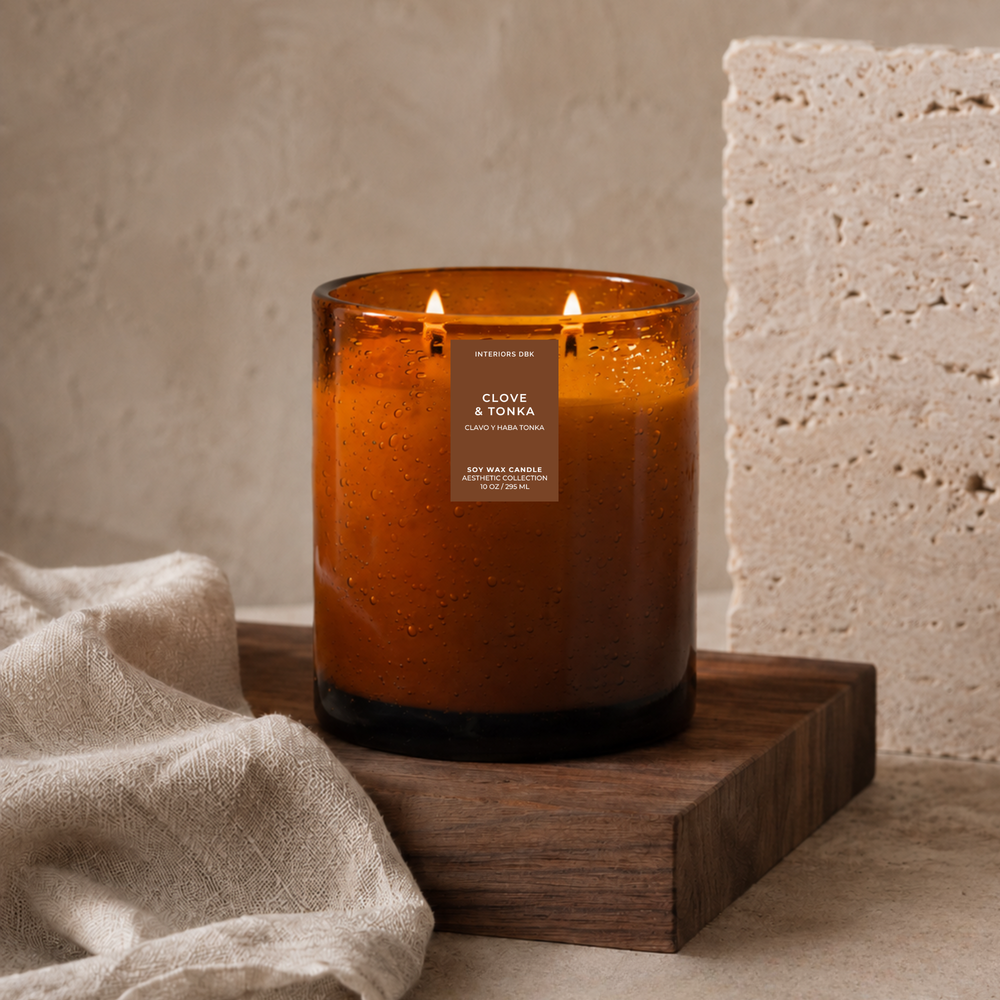 Clove and Tonka soy candle by Interiors DBK. An amber glass candle with two lit wicks sits on a dark wooden block, styled with a beige linen cloth and a textured stone block against a neutral beige background.