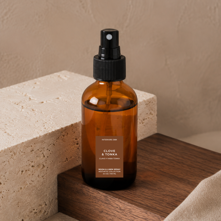 Clove and Tonka by Interiors DBK. An amber glass room and linen spray bottle with a black pump dispenser styled on a wooden surface next to a textured stone block.