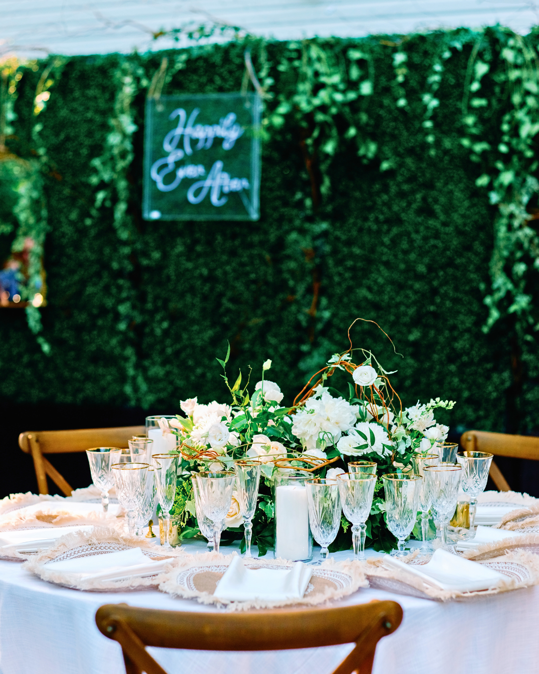 Luxury event design by Interiors DBK. Decorated table with white flowers and glasses against a greenery backdrop with 'Happily Ever After' sign.