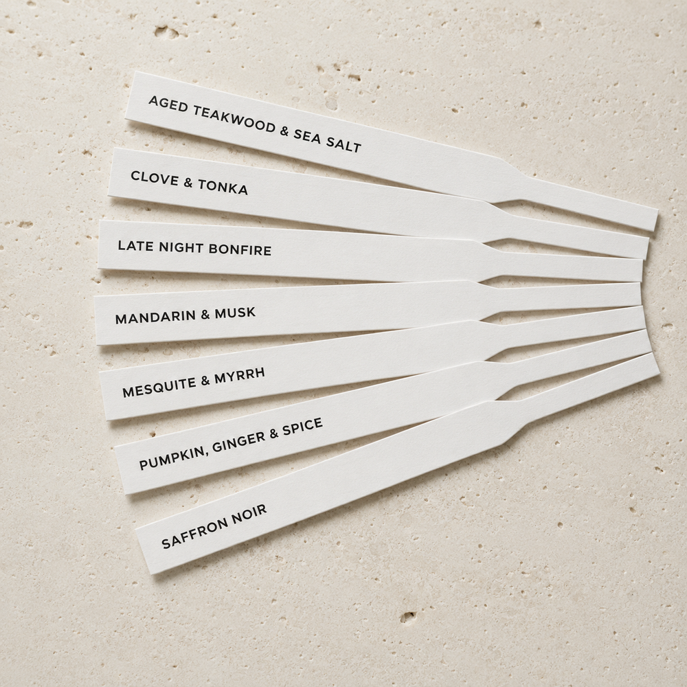 Seven white rectangular scent strips with black text listing different fragrance notes, arranged in a fan shape on a light beige textured surface. The notes include aged teakwood and sea salt, clove & tonka, late night bonfire, mandarin & musk, mesquite & myrrh, pumpkin, ginger & spice, and saffron noir.
