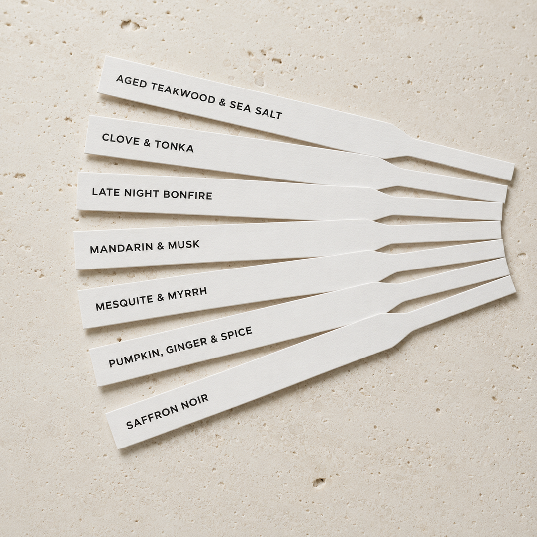 Seven white rectangular scent strips with black text listing different fragrance notes, arranged in a fan shape on a light beige textured surface. The notes include aged teakwood and sea salt, clove &amp; tonka, late night bonfire, mandarin &amp; musk, mesquite &amp; myrrh, pumpkin, ginger &amp; spice, and saffron noir.