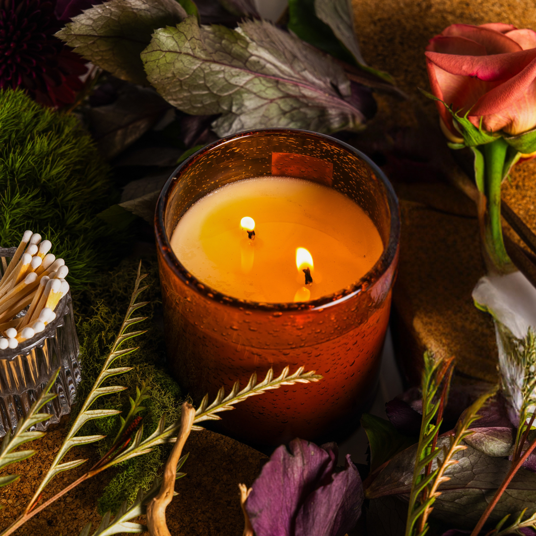 A lit amber glass candle with two wicks, surrounded by dried flowers, greenery, moss, and a small glass matchbox in a styled tabletop setting. Handcrafted by Interiors DBK