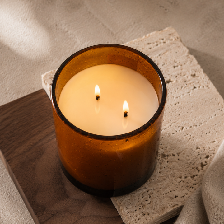Soy wax candle by Interiors DBK. A lit amber glass candle with two wicks sits on a wooden board and a textured stone tile against a neutral fabric background.