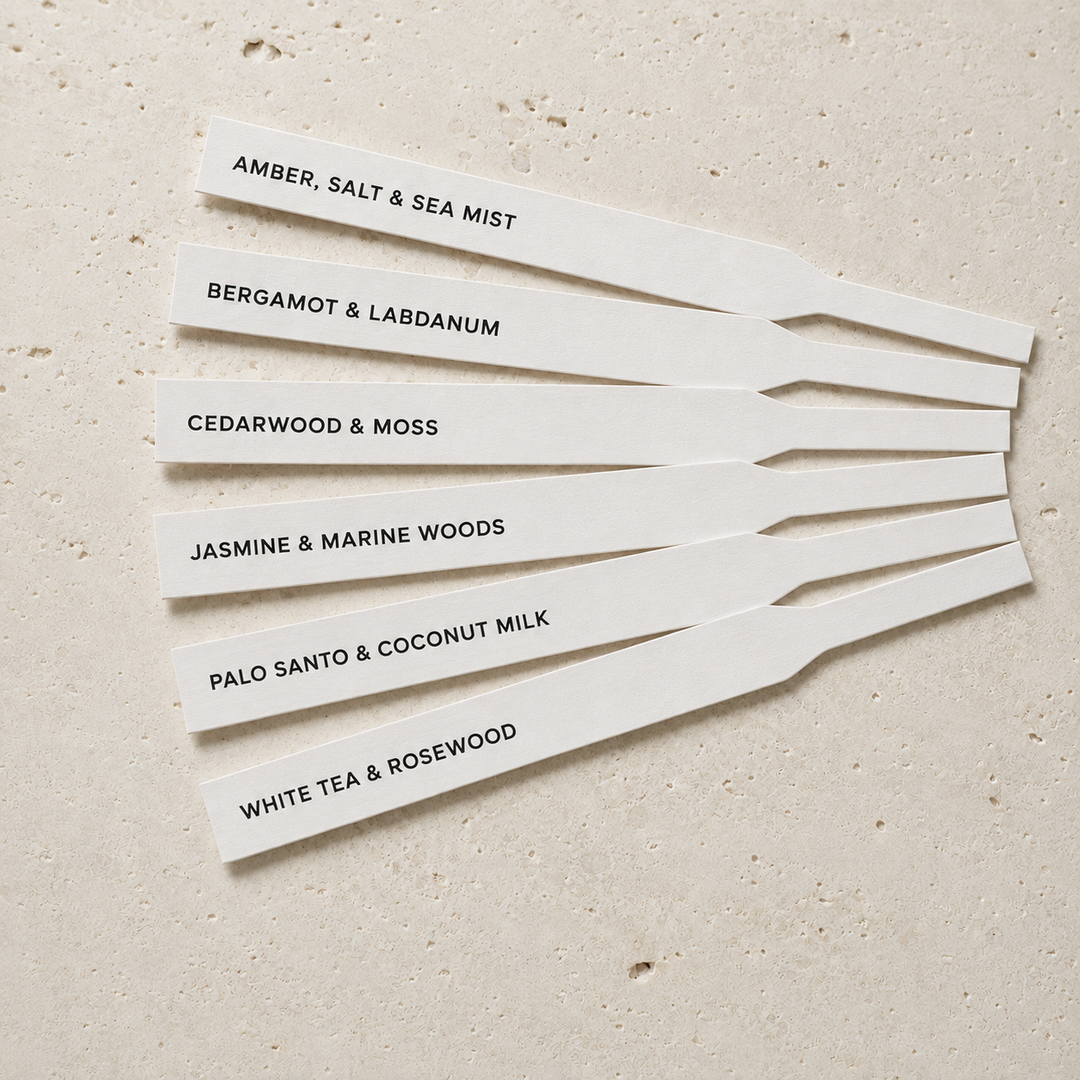 Six white rectangular scent cards with black printed text, arranged in a fan shape on a light beige textured surface. The cards read: AMBER, SALT &amp; SEA MIST, BERGAMOT &amp; LABDANUM, CEDARWOOD &amp; MOSS, JASMINE &amp; MARINE WOODS, PALO SANTO &amp; COCONUT MILK, and WHITE TEA &amp; ROSEWOOD.