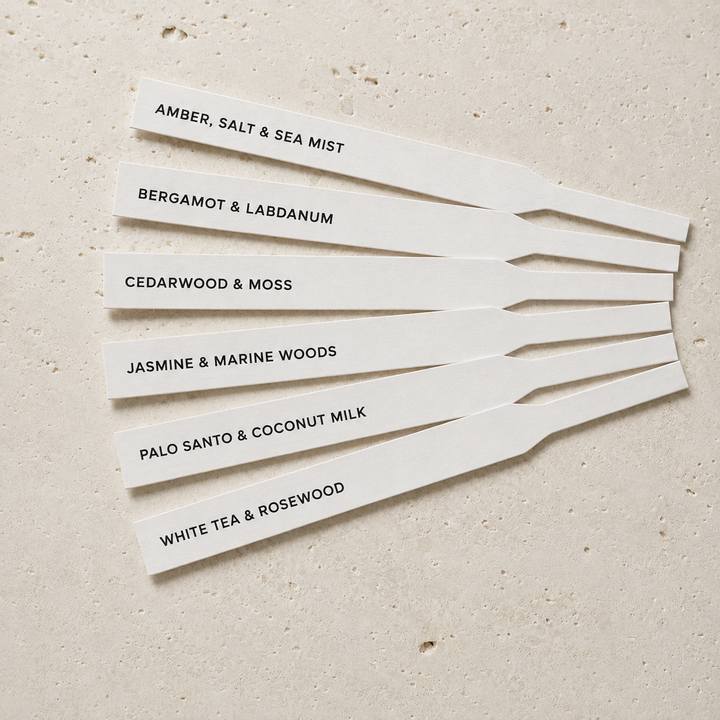 Six white rectangular scent cards with black printed text, arranged in a fan shape on a light beige textured surface. The cards read: AMBER, SALT &amp; SEA MIST, BERGAMOT &amp; LABDANUM, CEDARWOOD &amp; MOSS, JASMINE &amp; MARINE WOODS, PALO SANTO &amp; COCONUT MILK, and WHITE TEA &amp; ROSEWOOD.