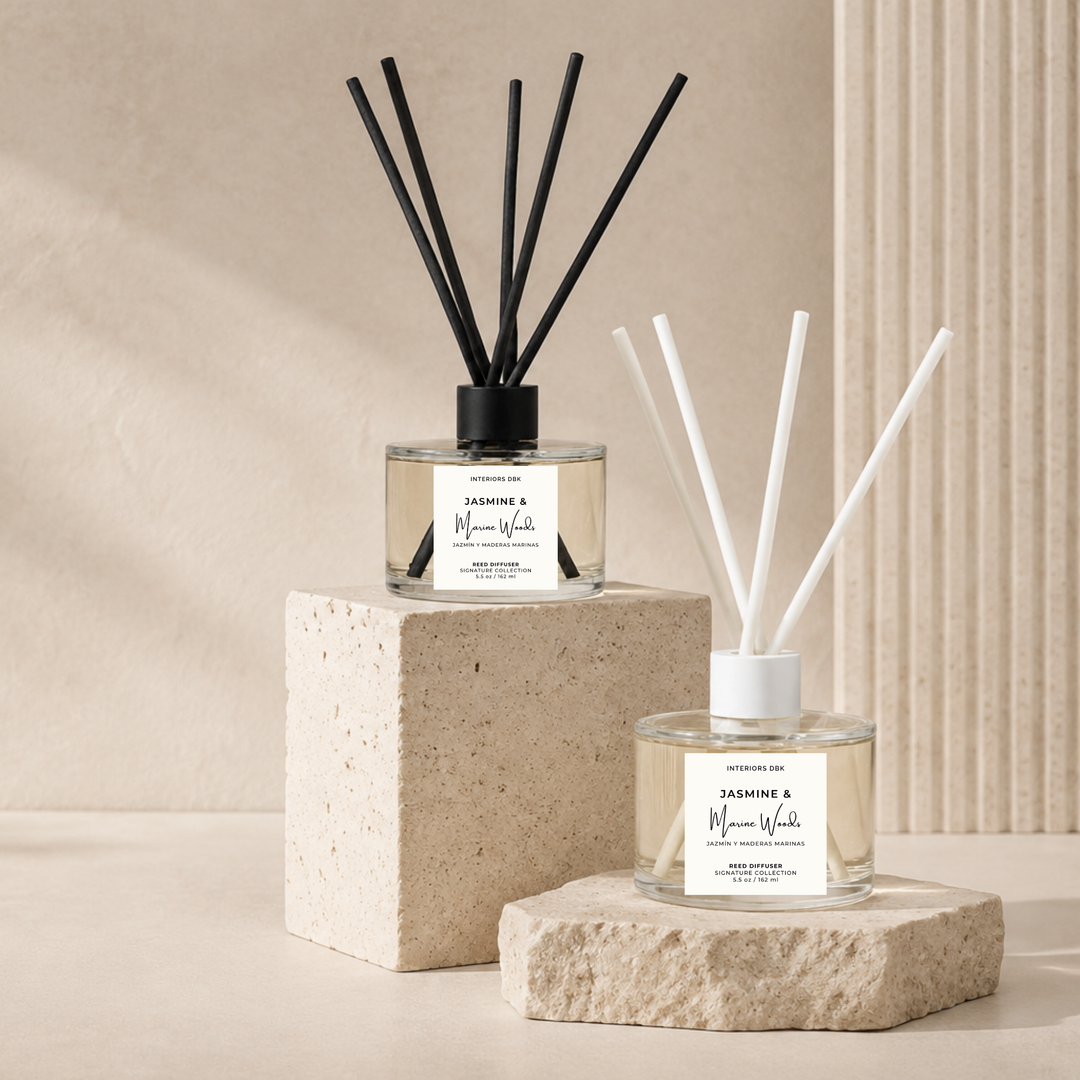 Two Jasmine and Marine Woods reed diffusers with clear glass bottles styled on stone blocks against a warm beige wall. One diffuser has black reeds and sits on a larger cube, while the other has white reeds and sits on a smaller rectangular block in front. 