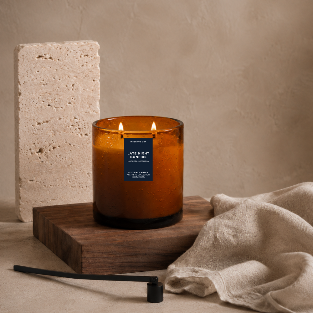Late Night Bonfire soy wax candle designed in an amber glass jar candle by Interiors DBK sits on a wooden block, styled with a linen cloth, a stone tile, and a black wick holder against a warm beige textured background.