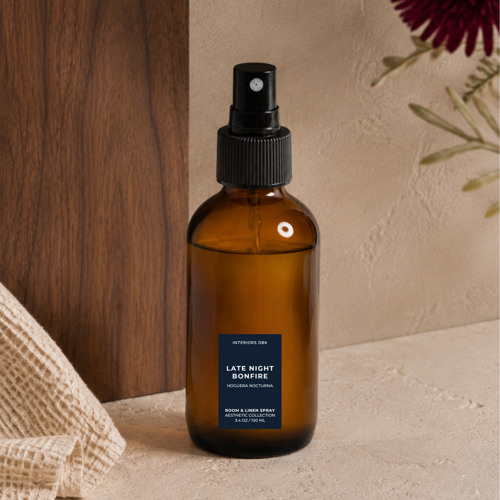 Late Night Bonfire room and linen spray in amber glass bottle, smoky woodsy home fragrance by Interiors DBK set on a textured beige surface next to a wooden panel and burlap fabric.