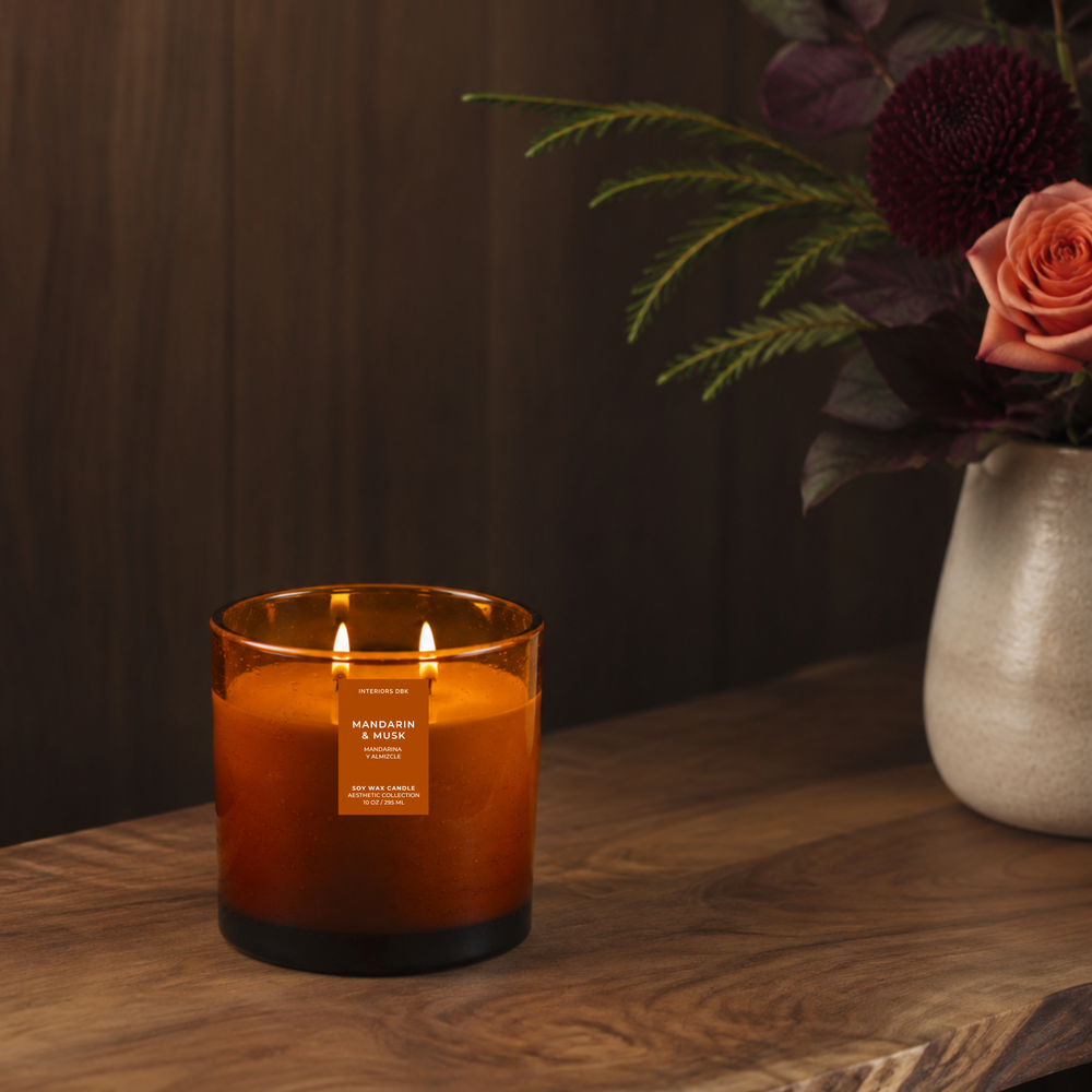 Mandarin and Musk soy wax candle by Interior DBK with two lit wicks sits on a wooden table next to a ceramic vase with flowers, against a dark wood-paneled background.