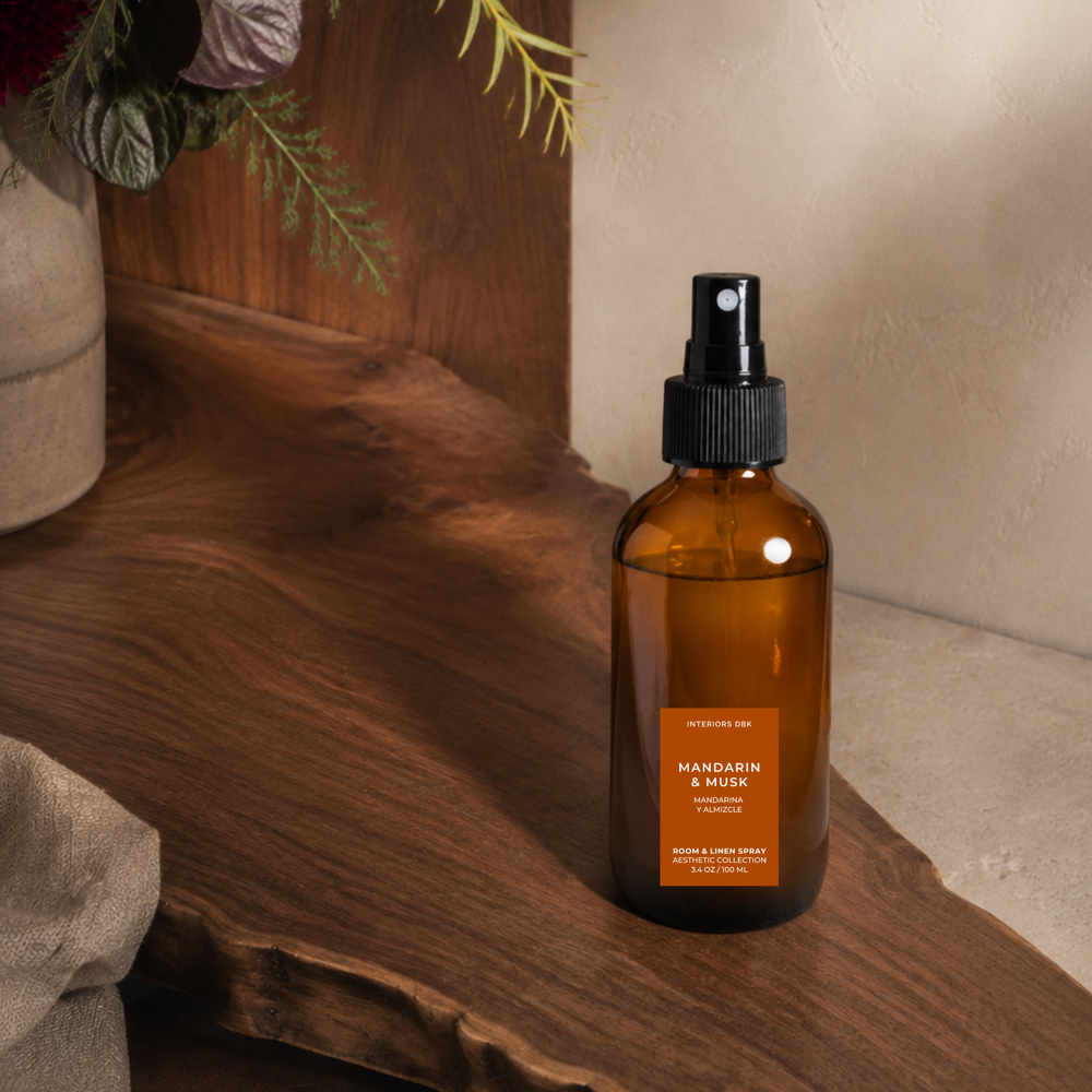 Mandarin and Musk room and linen spray in amber glass bottle, citrus wood home fragrance by Interiors DBK set on a rustic wooden slab with a neutral wall and dried flowers in the background.
