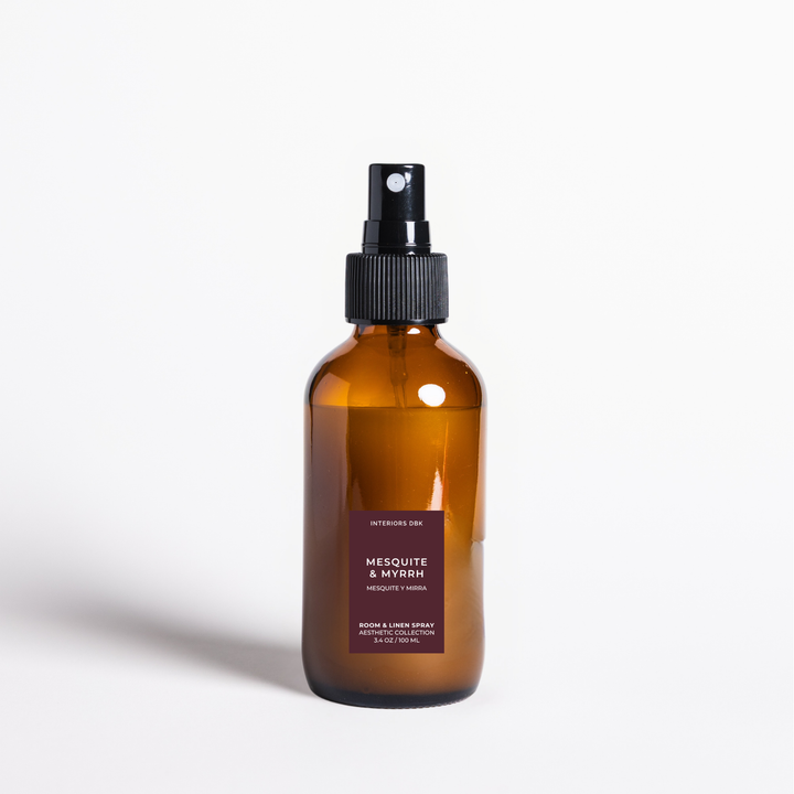 Mesquite and Myrrh by Interiors DBK. An amber glass room and linen spray bottle with a black pump dispenser photographed on a clean white background.