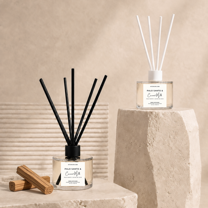 Two Palo Santo and Coconut Milk reed diffusers displayed on a textured stone pedestal and floor against a warm beige background. One diffuser on the left has black reeds and a black cap, resting next to two wooden blocks. Flameless and non-toxic aromas by Interiors DBK.