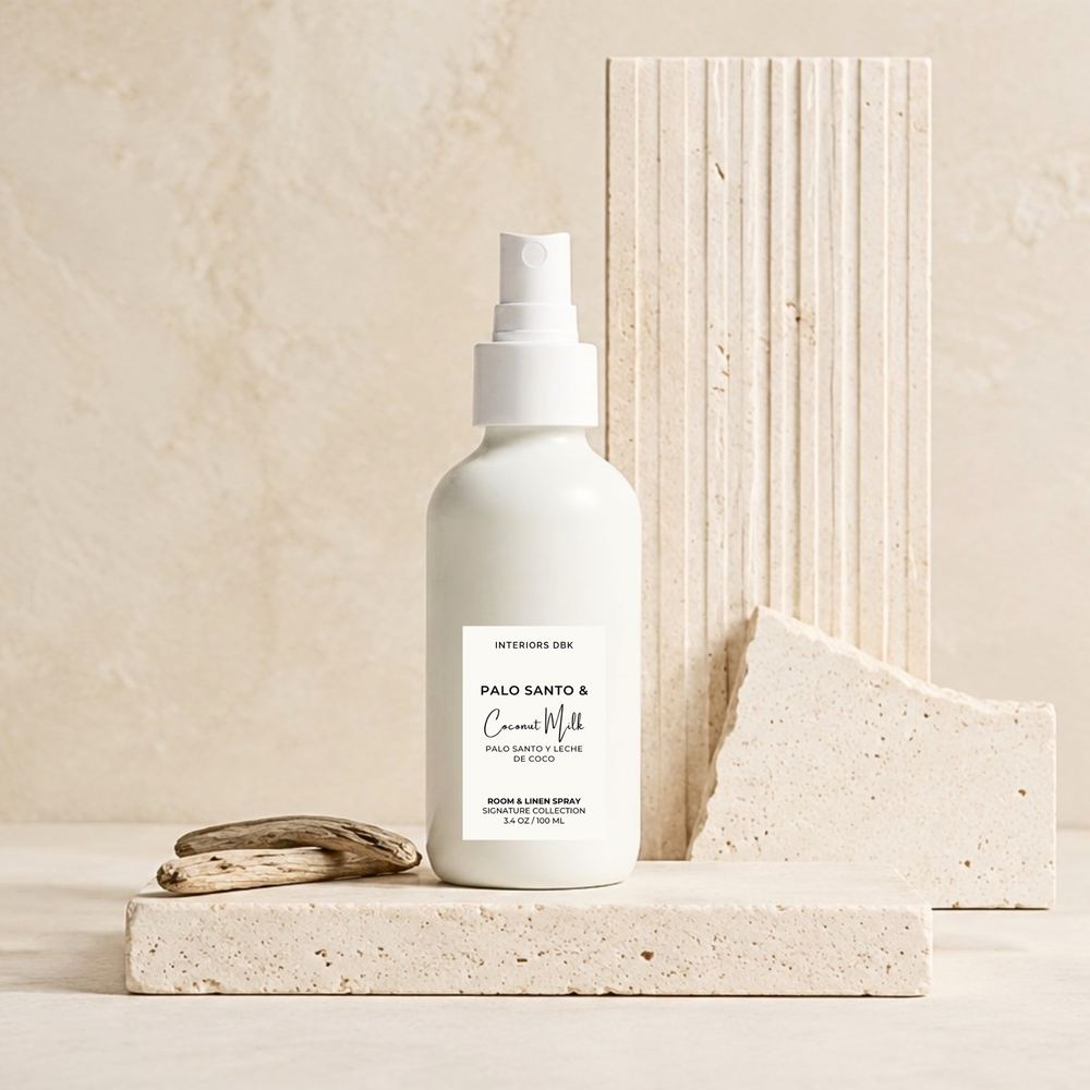 Palo Santo & Coconut Milk room spray by Interiors DBK in white glass bottle stands on a light stone platform, with a small piece of driftwood beside it and textured stone blocks behind it in a warm neutral setting.