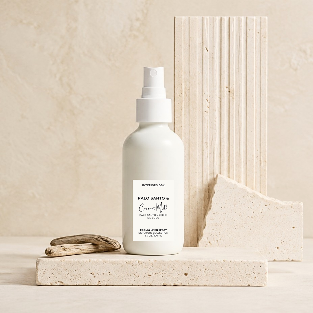 Palo Santo &amp; Coconut Milk room spray by Interiors DBK in white glass bottle stands on a light stone platform, with a small piece of driftwood beside it and textured stone blocks behind it in a warm neutral setting.
