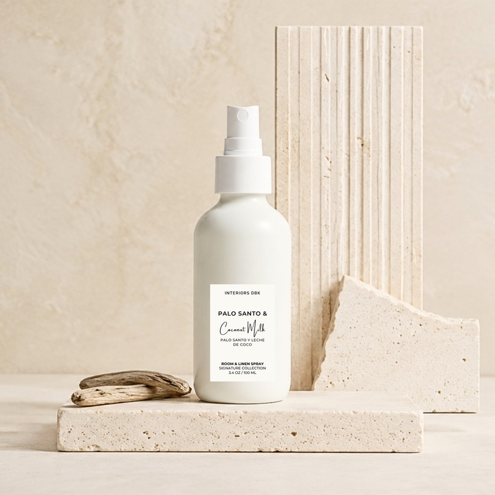 Palo Santo &amp; Coconut Milk room spray by Interiors DBK in white glass bottle stands on a light stone platform, with a small piece of driftwood beside it and textured stone blocks behind it in a warm neutral setting.