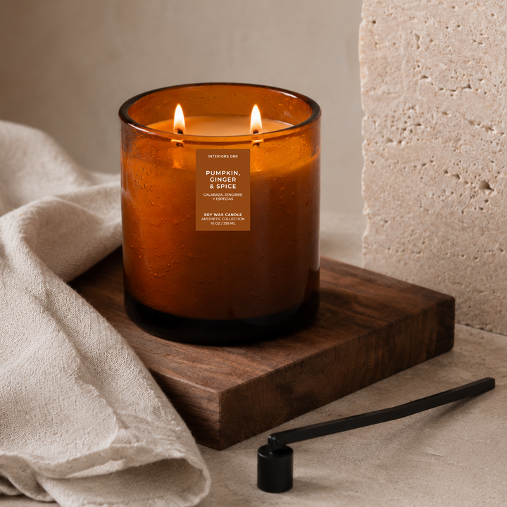 Pumpkin, Ginger and Spice soy wax candle handmade by Interiors DBK. A lit amber glass candle with two flames sits on a dark wooden block, with a beige cloth draped to the left and a black candle snuffer nearby on a neutral textured surface.