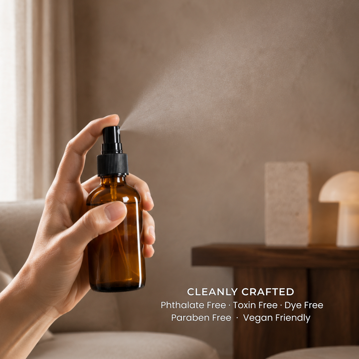 Room and Linen Sprays by Interiors DBK. Designed in reusable amber glass spray bottles with a black pump dispenser. A hand holds a small amber glass spray bottle with a black pump, releasing a fine mist in a softly lit indoor setting. Text on the image reads: &