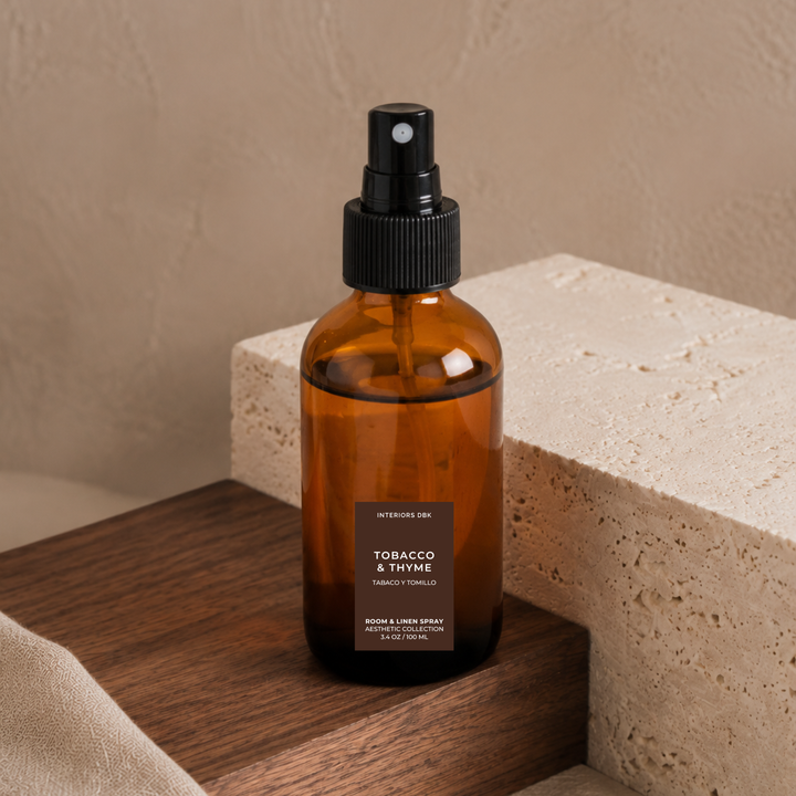 Tobacco and Thyme by Interiors DBK. An amber glass room and linen spray bottle with a black pump dispenser. Styled on a wooden block next to a textured stone block against a beige wall.